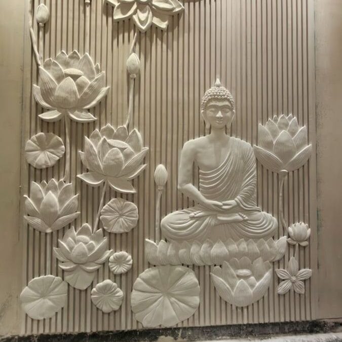 3D white relief mural of a serene Buddha seated in meditation, surrounded by large lotus flowers and lily pads, set against a vertically grooved textured wall with soft overhead lighting.