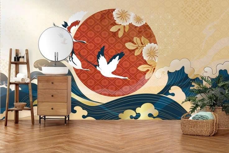 A bathroom interior with a large wall mural featuring three white cranes flying across a bold red sun circle adorned with floral patterns. Large, stylized blue and gold ocean waves sweep across the bottom of the mural against a light beige background with geometric motifs. A wooden vanity and round mirror sit to the left.