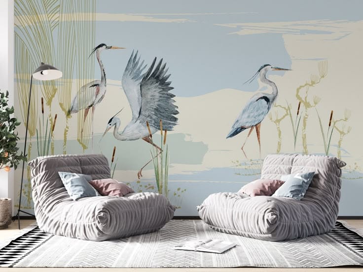 A peel-and-stick wallpaper mural featuring watercolor-style herons or cranes standing in a stylized marsh landscape with cattails, set behind two plush, modern grey lounge chairs and a patterned area rug.