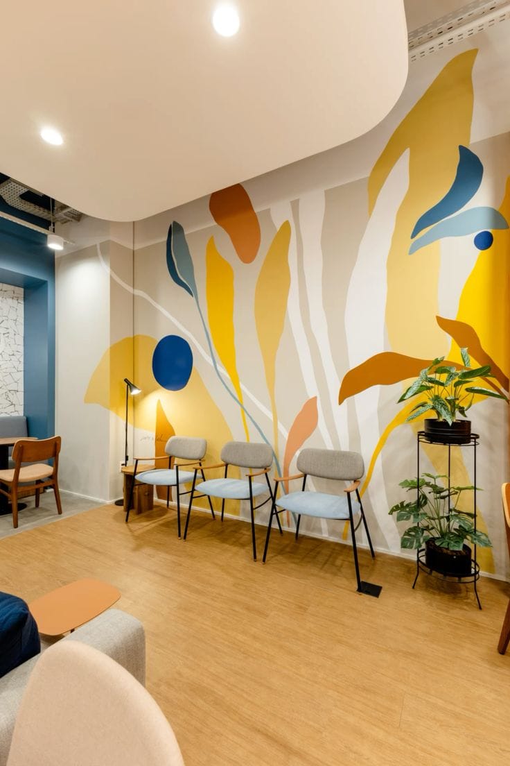 A well-lit contemporary waiting room featuring a large, colorful abstract mural of organic leaf shapes in yellow, blue, and orange. Three light blue chairs with wooden armrests are lined up against the wall, complemented by light wood flooring and indoor plants on a black metal stand.