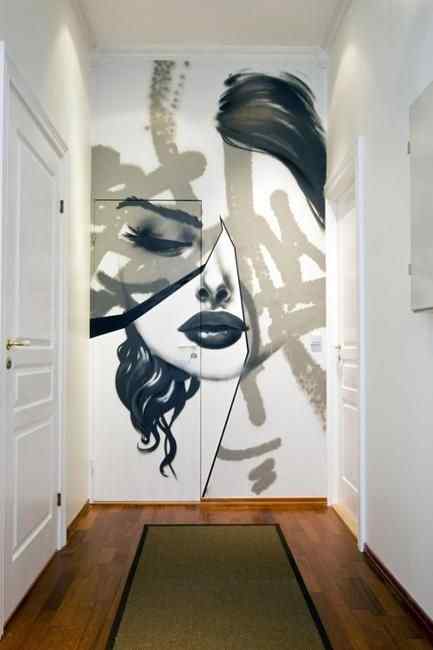 A monochrome mural at the end of a white hallway featuring a woman's face. The portrait is "fractured" or split by geometric lines and overlaid with abstract, grey graffiti-style brushstrokes.