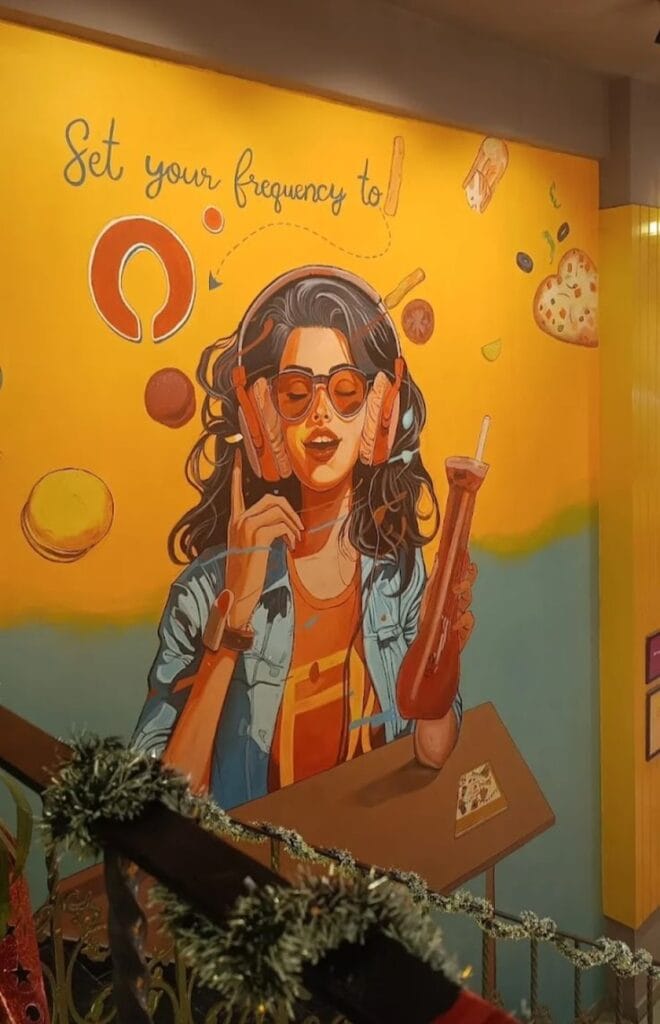 A warm, yellow-toned cafe mural featuring an illustration of a woman with long dark hair wearing large headphones and orange sunglasses. She is holding a tall drink. Surrounding her are floating food icons like a burger and pizza. The text above reads, "Set your frequency to."