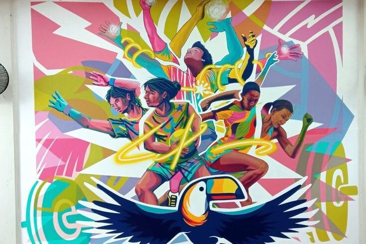 A square indoor mural on a white wall. At the bottom center is a stylized blue and orange toucan with outstretched wings. Above it, several athletes—including a weightlifter and basketball players—are depicted in a colorful, overlapping style with glowing yellow swirls surrounding them.