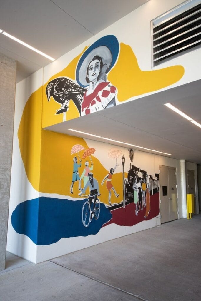 A large mural in a hallway featuring bold yellow, blue, and maroon organic shapes layered with black-and-white vintage illustrations of a woman, a cyclist, and a steam train.