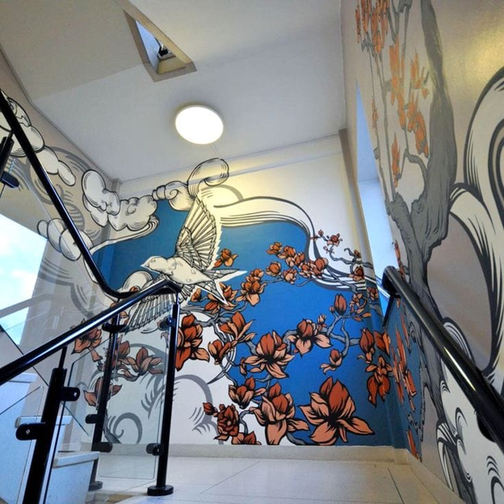 A two-story stairwell featuring a wrap-around mural of a white bird in flight against a bright blue background. The mural includes large, stylized orange blossoms on grey branches and flowing white cloud patterns.