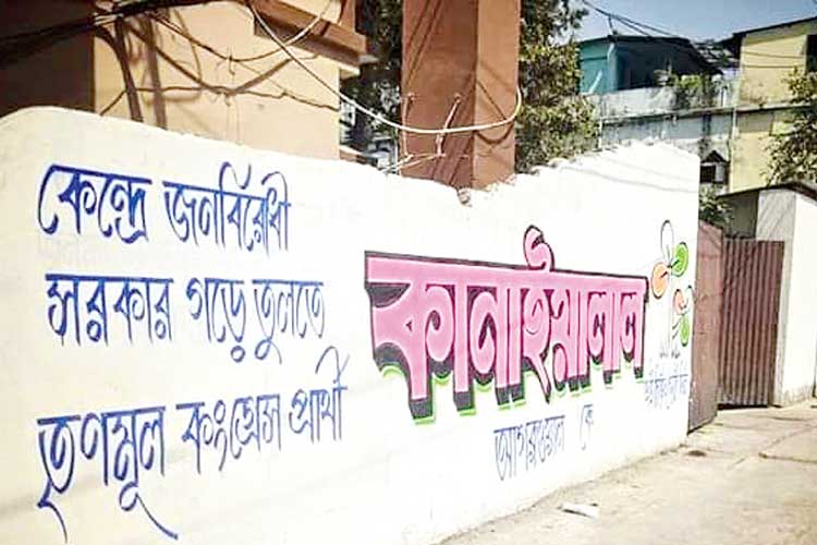 A long white boundary wall featuring hand-painted Bengali calligraphy in blue and pink, representing local political campaigning and traditional sign-painting art.