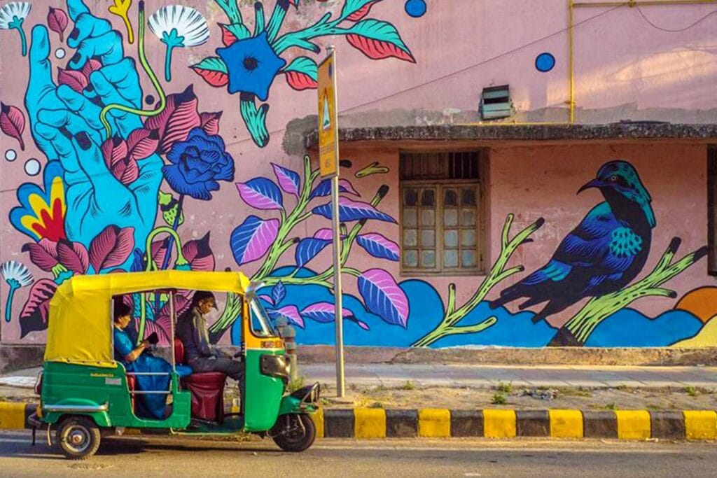 A colorful outdoor wall mural featuring large blue flowers, pink leaves, and a bird, with a green and yellow auto-rickshaw passing by on a city street.