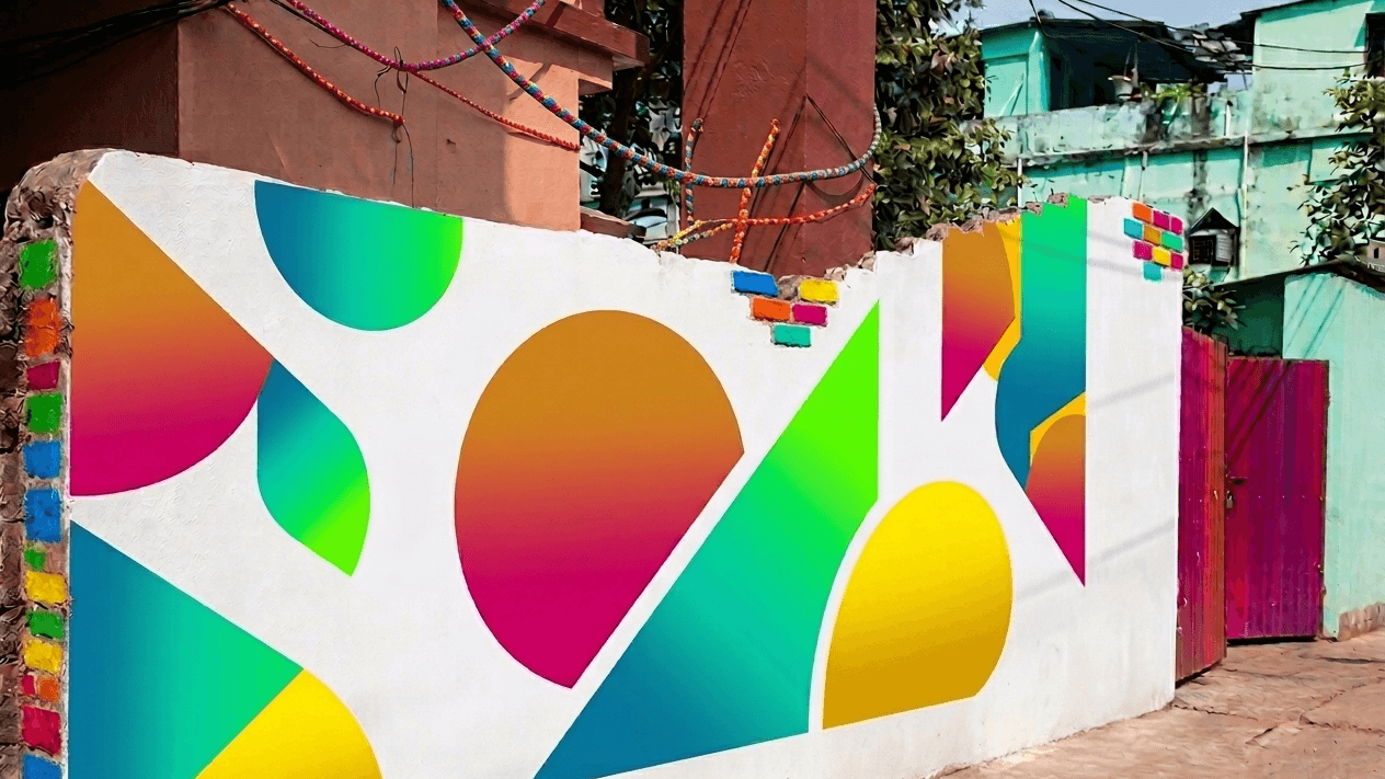 An outdoor, eye-level shot of a white wall featuring a vibrant, abstract graffiti mural. The mural is composed of large, geometric shapes—including circles, triangles, and semi-circles—filled with bright, multi-colored gradients of pink, orange, yellow, green, and blue. To the left, the edge of the wall is decorated with a vertical stack of colorful, painted bricks. The wall has a rough, uneven top edge and is located in an alleyway next to a red gate and a light green building. Above the wall, tangled power lines are visible against a clear, pale sky. The ground is a dusty, unpaved path.