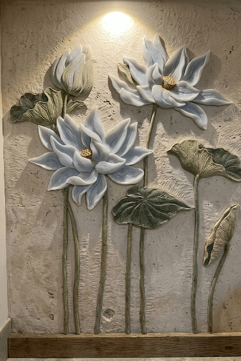A vertical wall-mounted relief sculpture featuring three-dimensional lotus flowers and large green leaves on a textured, sand-colored stone background. The lotuses are pale white with subtle blue shading, and a warm spotlight shines down from the top, casting soft shadows that emphasize the depth of the carving.