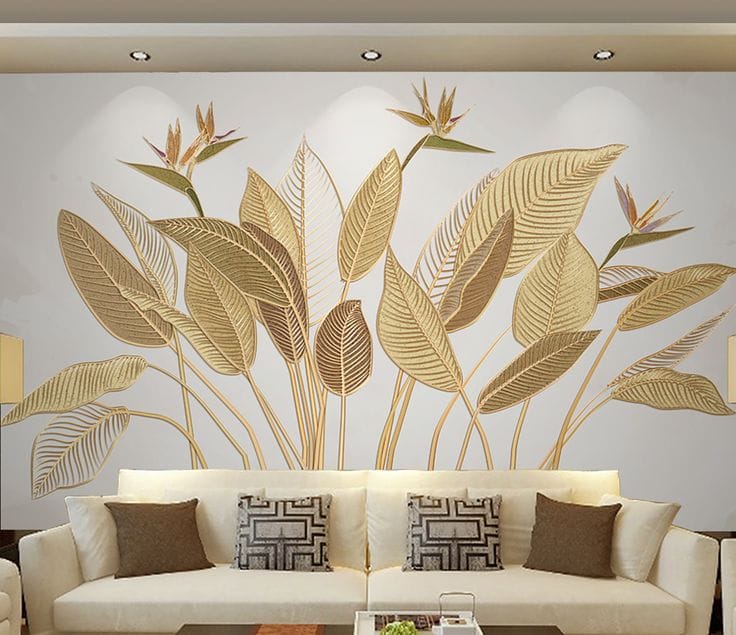 A wide-angle shot of a modern living room featuring a large-scale wall mural of stylized tropical leaves and Bird of Paradise flowers in gold and bronze tones. The mural is positioned behind a plush white contemporary sofa with decorative cushions, illuminated by recessed ceiling lights.
