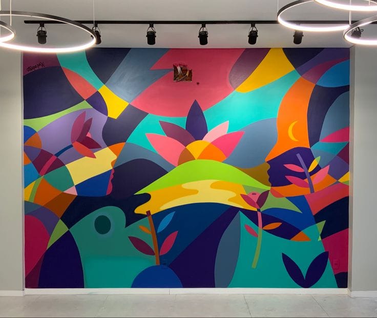 A large indoor wall mural featuring a colorful, abstract geometric design with stylized lotus flowers, leaves, and profiles of human faces in shades of pink, teal, orange, and purple.