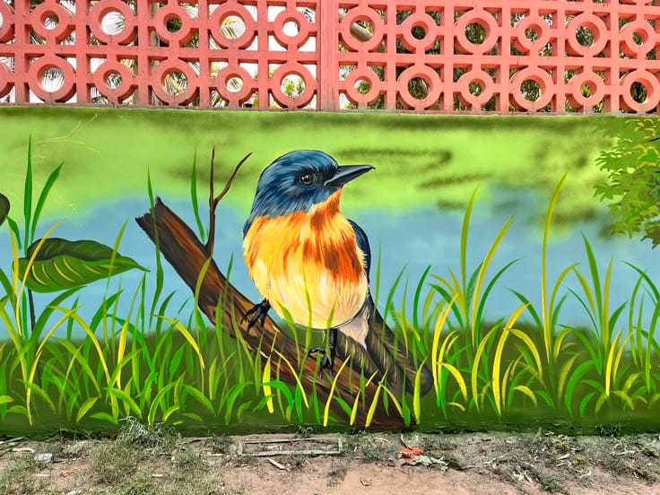 A colorful outdoor wall mural of a blue and orange songbird perched on a branch, surrounded by lush green grass and a soft blue sky.