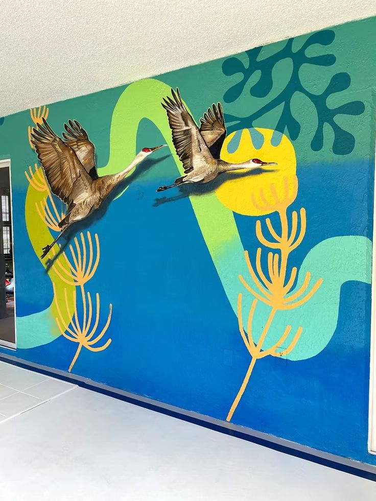 This large-scale outdoor mural features a central portrait of a woman with an upward gaze, framed by a vibrant yellow sun. To her left, two swallows are depicted in mid-flight, while to her right, a kingfisher rests on a branch. The background uses a striking diagonal split of sky blue and deep cerulean, creating a dynamic sense of movement across the long horizontal wall.