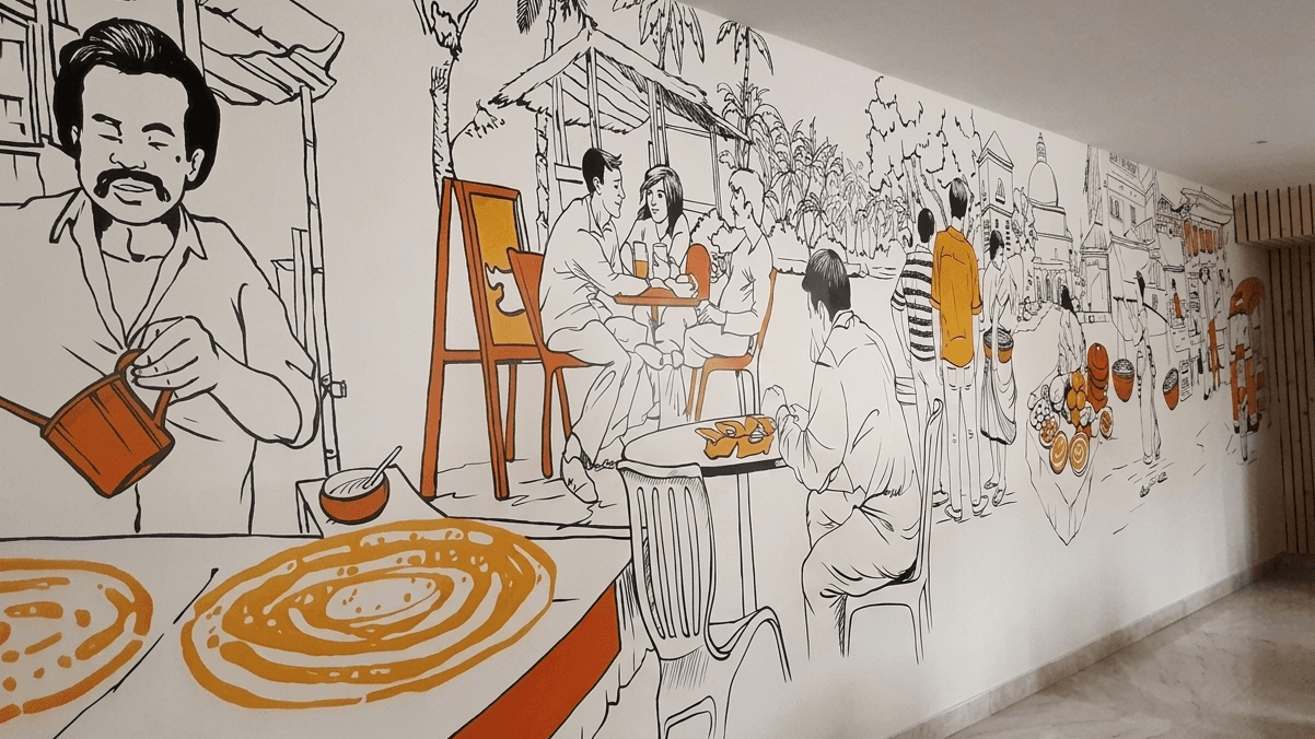 A wide-angle view of a long, indoor wall featuring a black and orange line-art mural. The artwork depicts various scenes of daily life and street food culture, including a man preparing food in the foreground, people dining at tables, and figures walking through a town with traditional architecture.