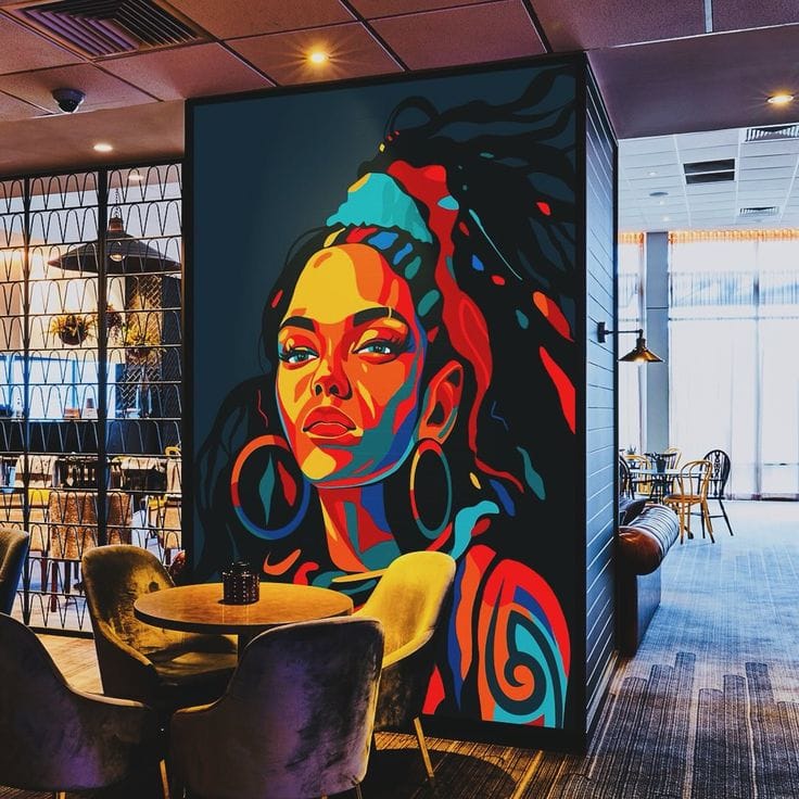 A high-contrast, colorful mural of a woman with a headband and large hoop earrings displayed on a dark pillar in a modern lounge or restaurant. The mural features a bold, pop-art style with shades of teal, red, and yellow. In the foreground, there are plush grey velvet chairs and a small round wooden table, set against a backdrop of a bright, contemporary interior with floor-to-ceiling windows and decorative metal partitions.