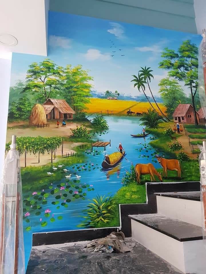 A detailed, floor-to-ceiling landscape mural painted on an interior wall next to a staircase. The artwork depicts a lush, tropical village scene with a winding blue river filled with lily pads and small boats. On the banks, there are thatched-roof huts, cows grazing, and villagers engaged in daily activities amidst golden fields and dense green trees. The sky is a clear light blue with small birds flying in the distance, creating an atmosphere of peace and nature.
