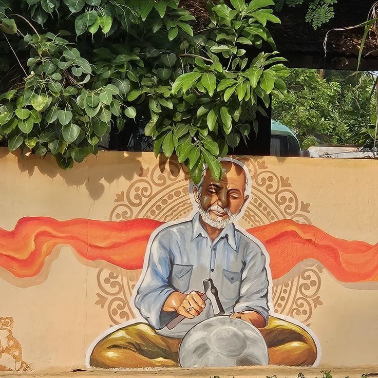 A vibrant outdoor mural on a tan wall featuring an elderly artisan with a white beard, meticulously hammering a metal bowl. Behind the craftsman is an intricate circular mandala pattern and a flowing orange ribbon-like graphic. Real green foliage from a tree hangs over the top of the wall, blending naturally with the painted scene. The artwork uses a bold, illustrative style with clean white outlines around the central figure.